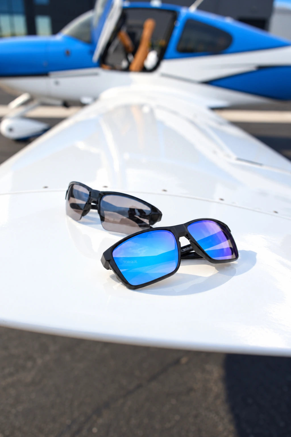 Blue mirrored and smoke-lens Torque sunglasses on a sunlit aircraft wing in front of a blue-and-white aircraft.