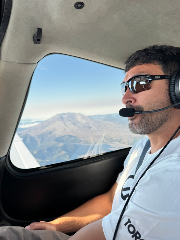 Pilot wearing sunglasses and a headset beside an aircraft window overlooking a mountain.