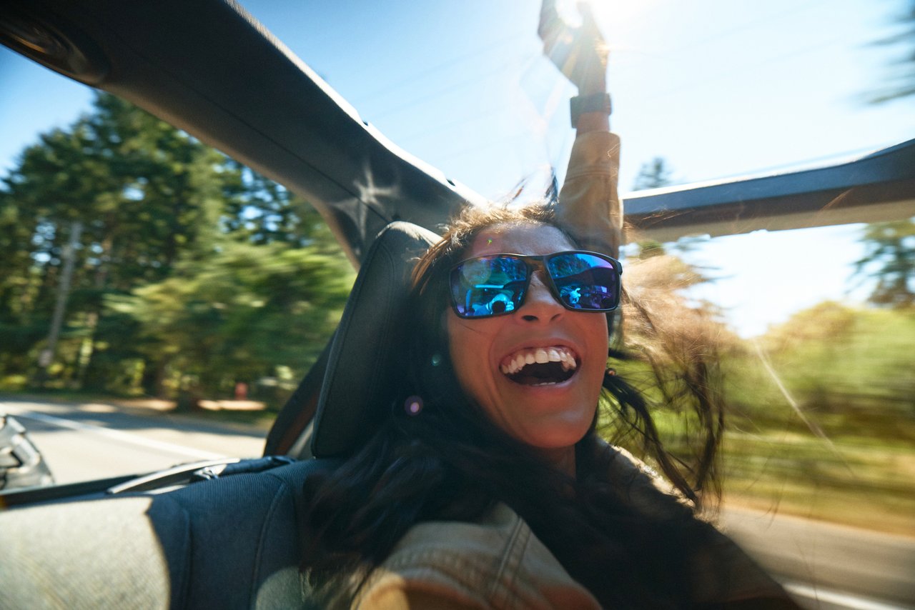 Smiling woman wearing Fulcrum sunglasses with blue mirrored lenses in a convertible