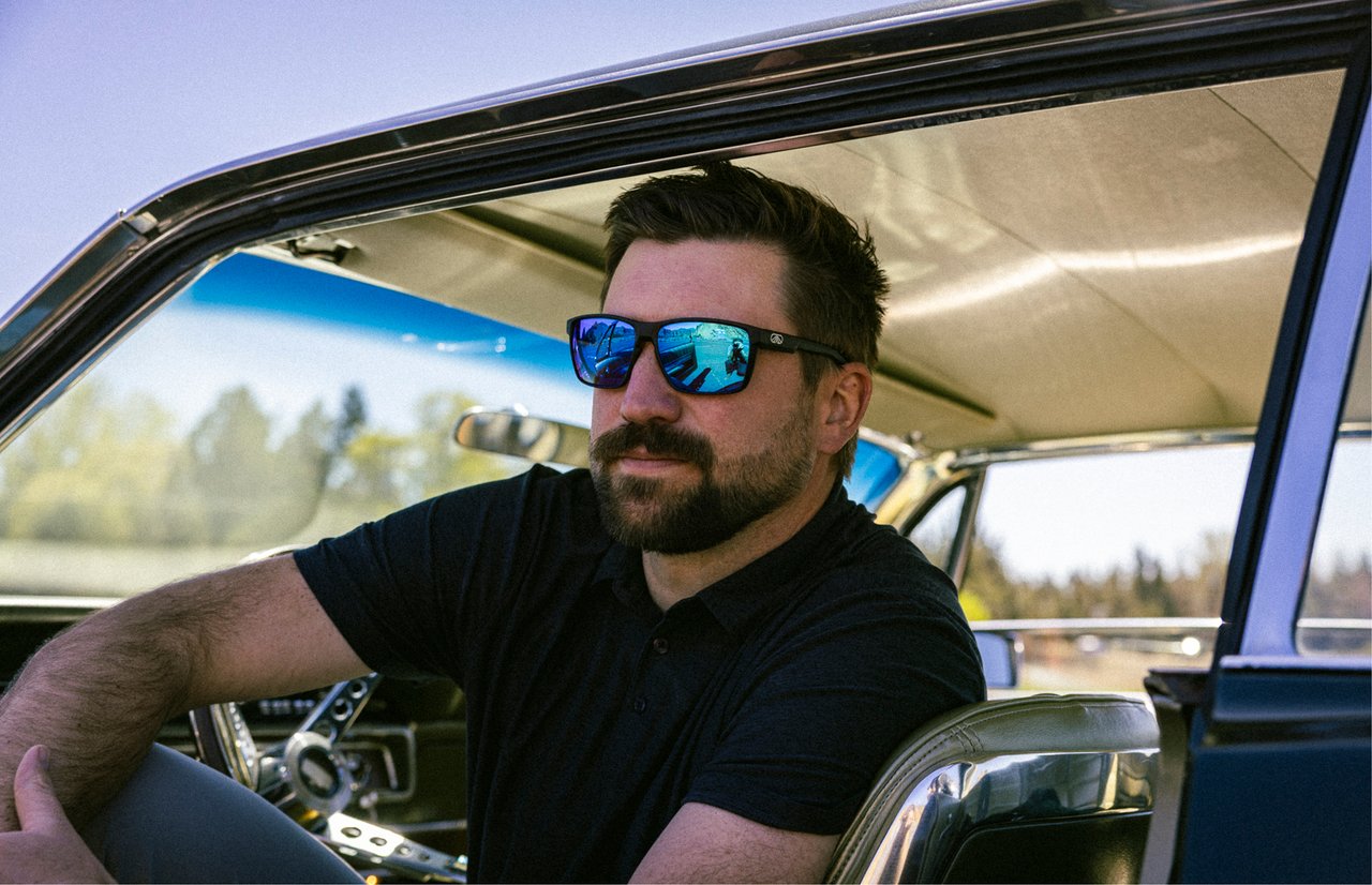 Man wearing black Fulcrum sunglasses with blue mirrored lenses in a classic car