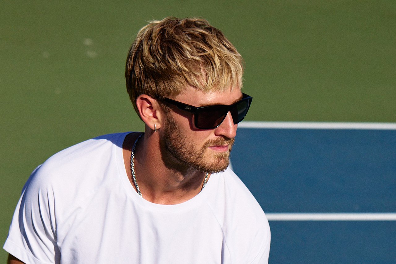 Man wearing black Fulcrum sunglasses and a white shirt on a court